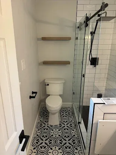 Small bathroom with toilet, patterned floor tiles, wall shelves, and glass shower enclosure.