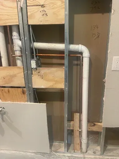 Exposed plumbing and wood framing in an unfinished wall with a white pipe bend and gray paneling.