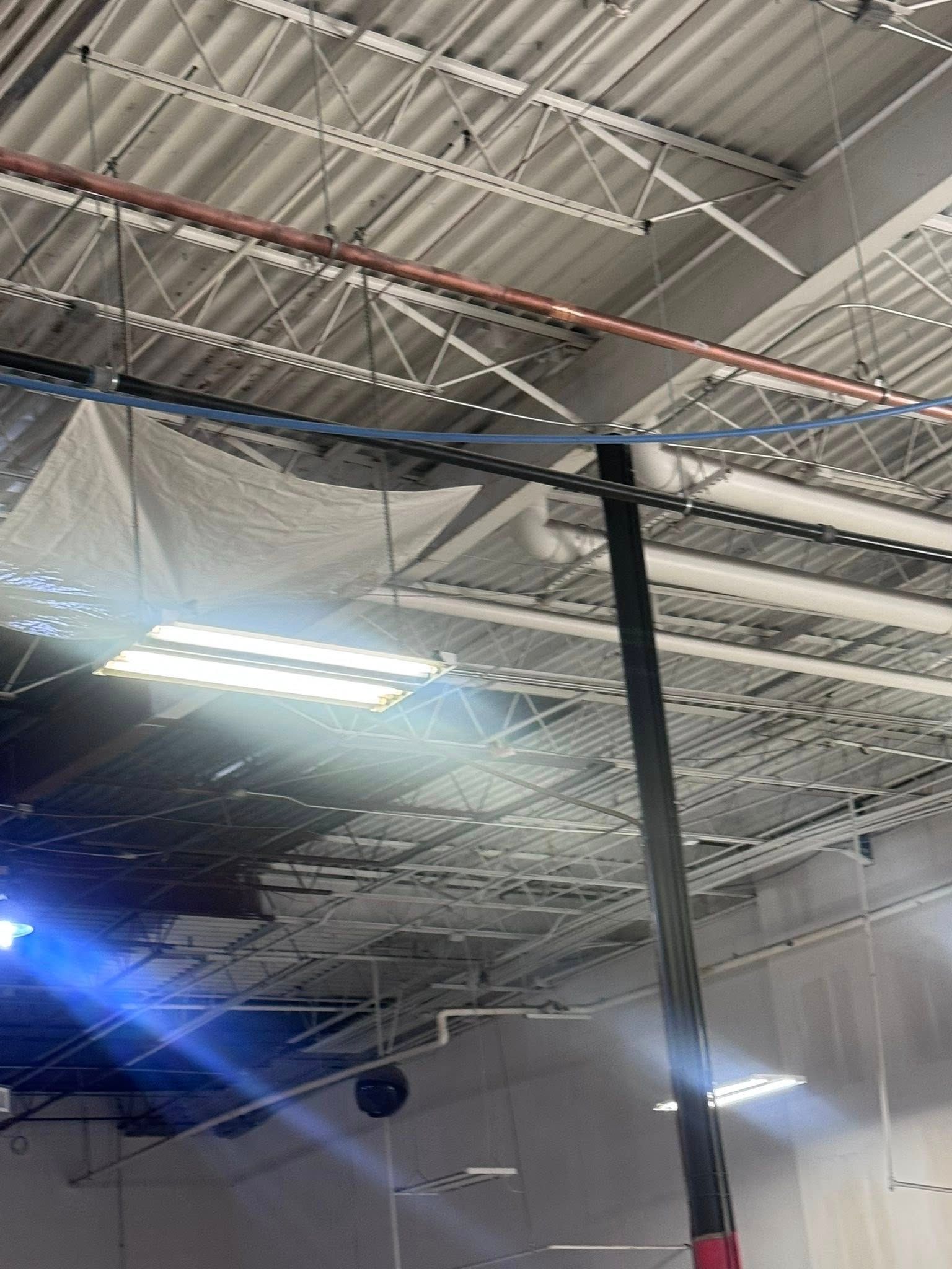 Industrial ceiling with exposed beams, hanging plastic sheeting, and bright overhead lights inside a warehouse.