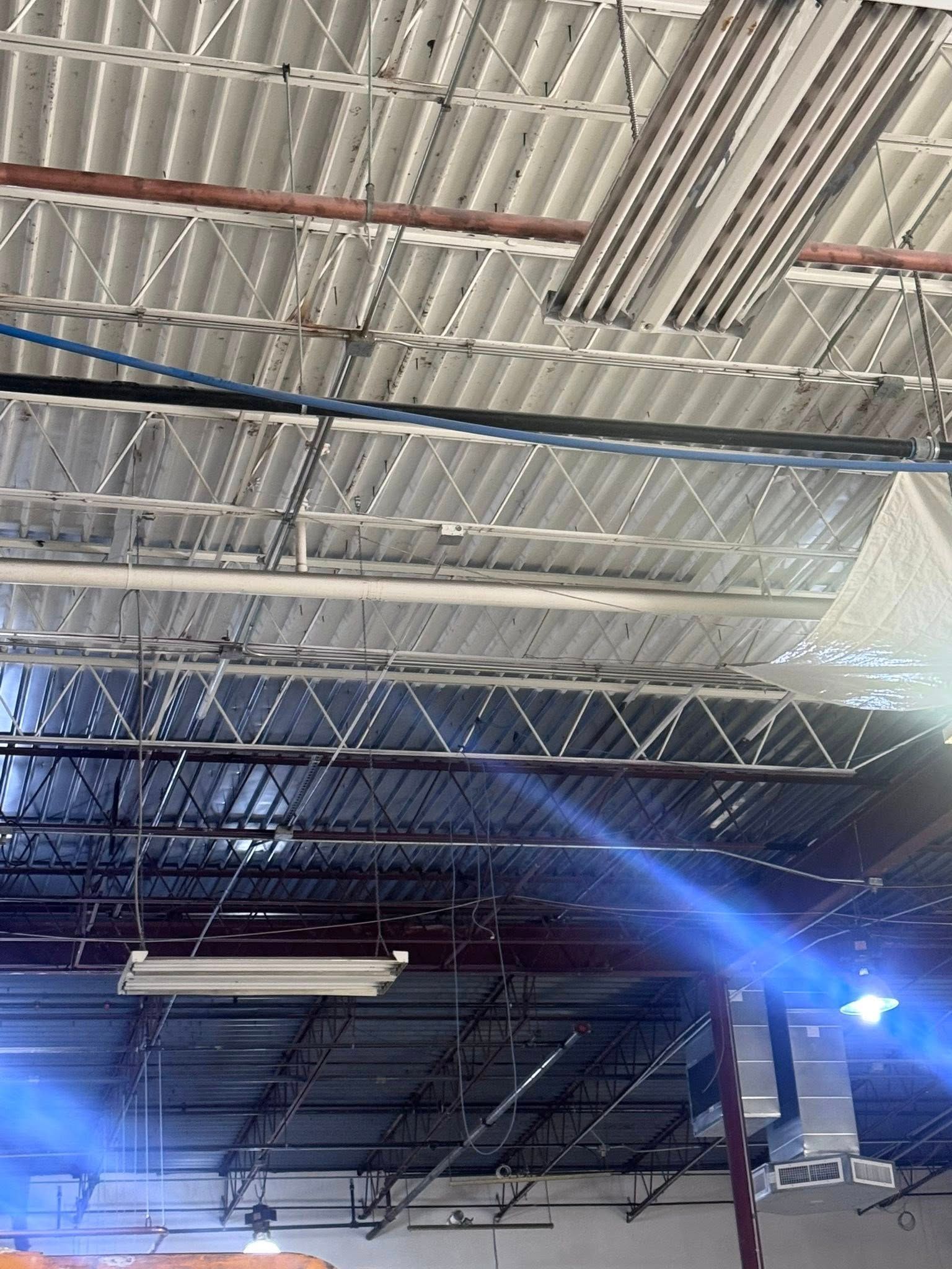 Indoor industrial ceiling with exposed metal beams, ducts, fluorescent lights, and blue stage lighting.