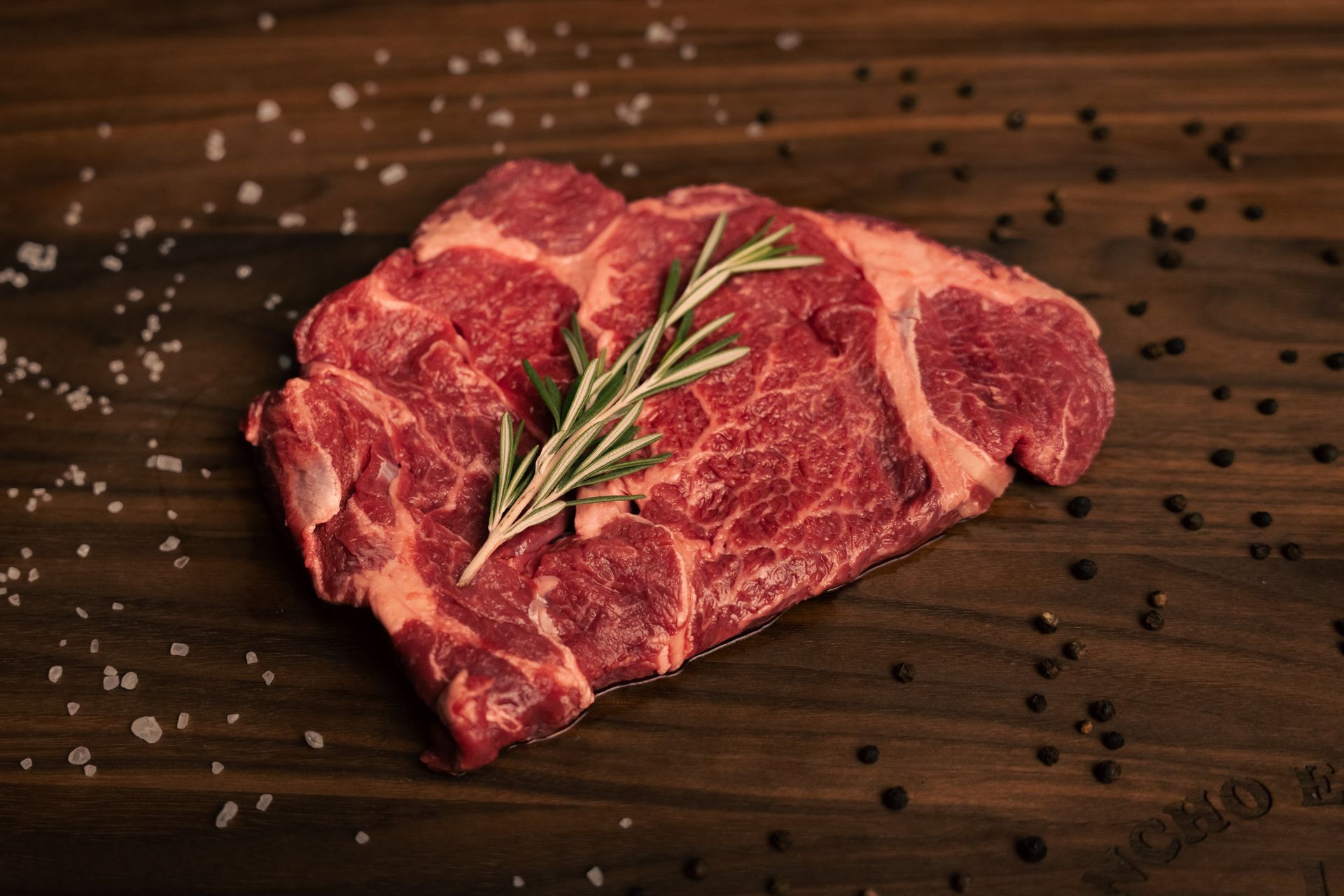 A piece of raw steak with rosemary on a wooden cutting board.