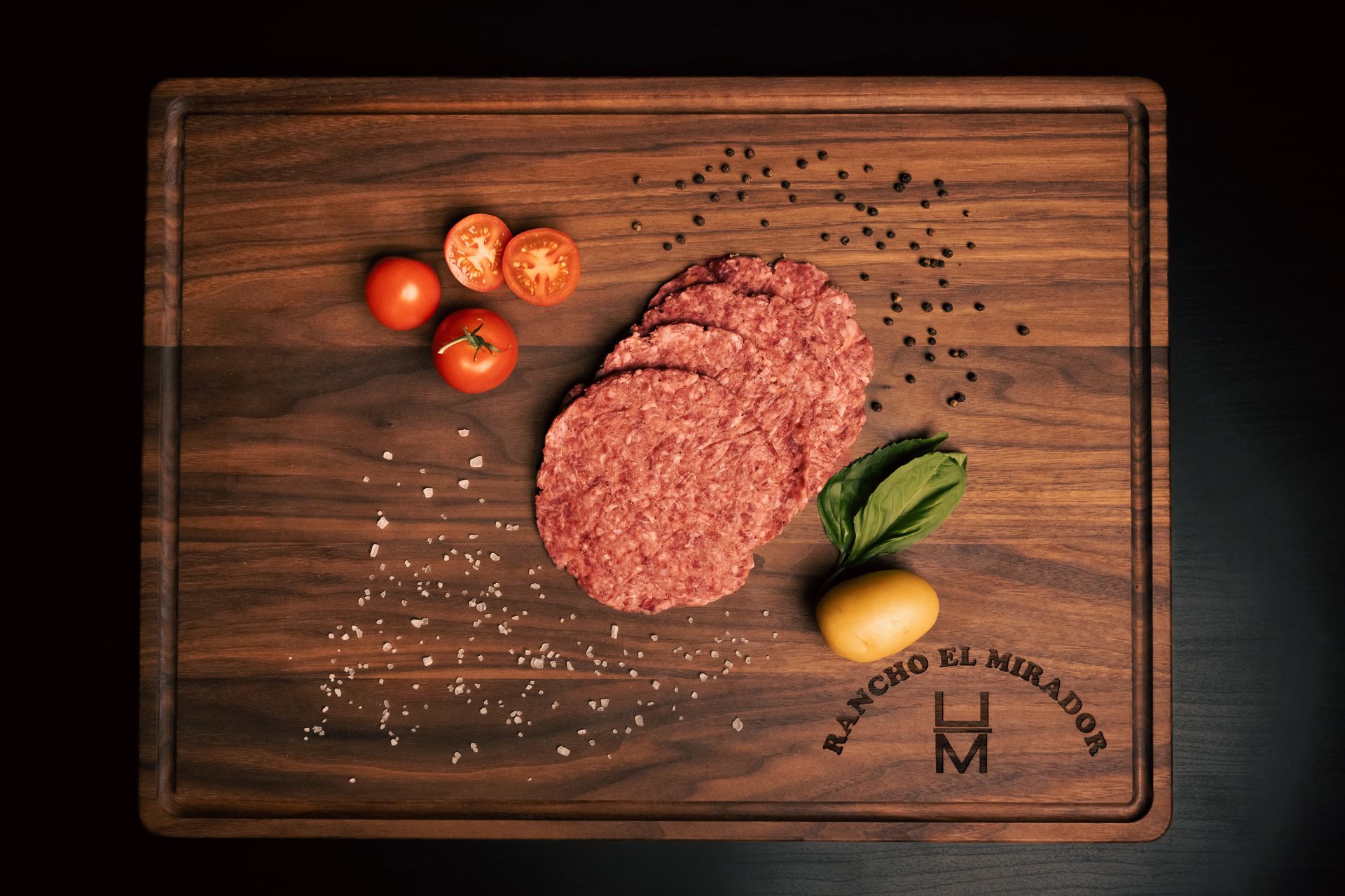 A wooden cutting board with hamburger patties and tomatoes on it.