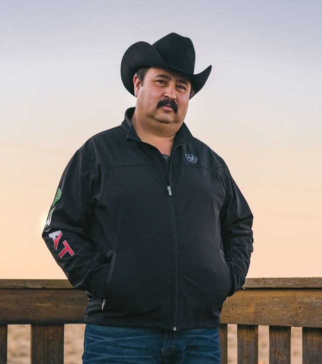 A man wearing a cowboy hat and a black jacket is standing next to a wooden railing.