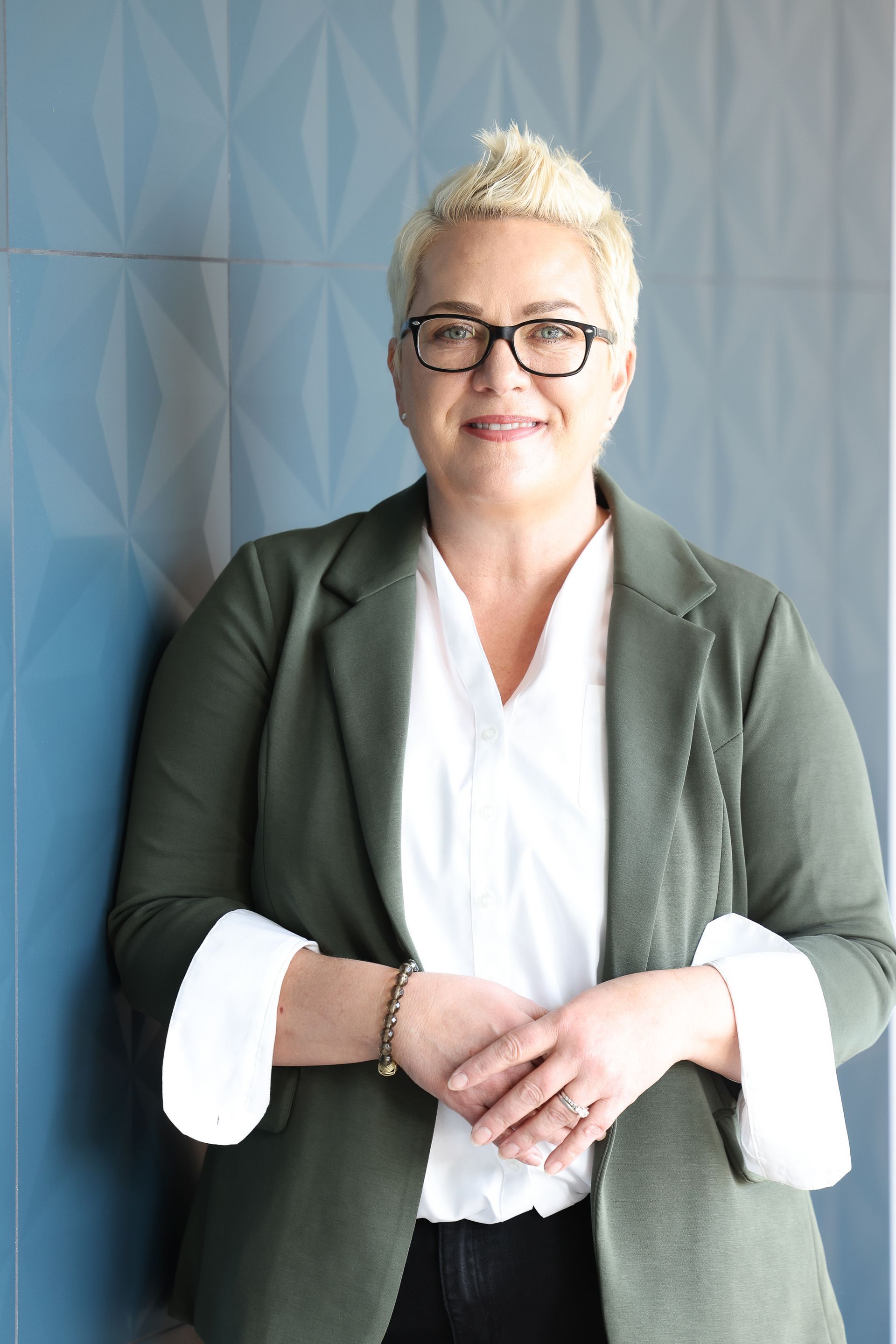 Person in glasses and green blazer standing against a gray textured wall, smiling with hands clasped.