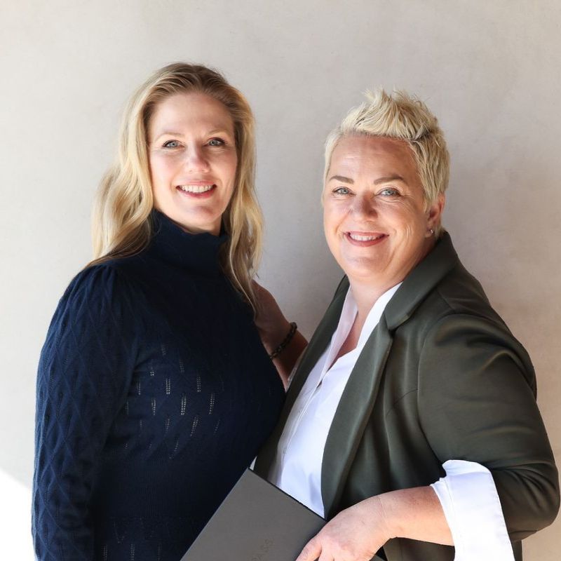Two smiling professionals standing side by side against a plain wall, holding folders