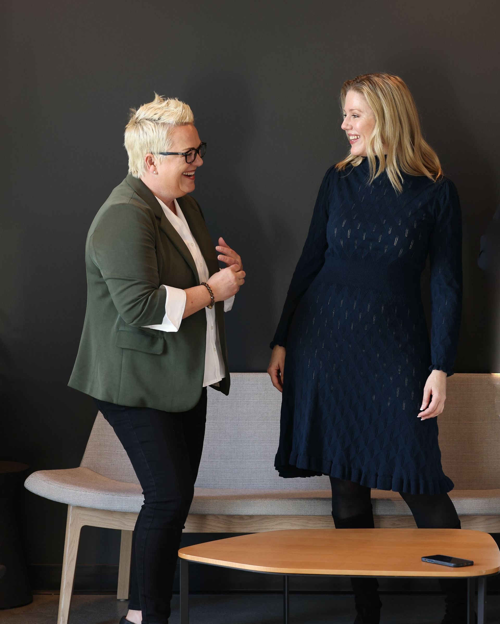 Two people standing and talking in a room beside a chair and round table.