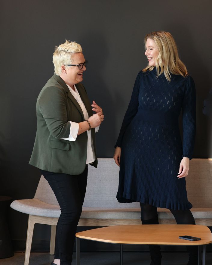 Two people smiling in conversation indoors, one standing and one beside a chair and table.