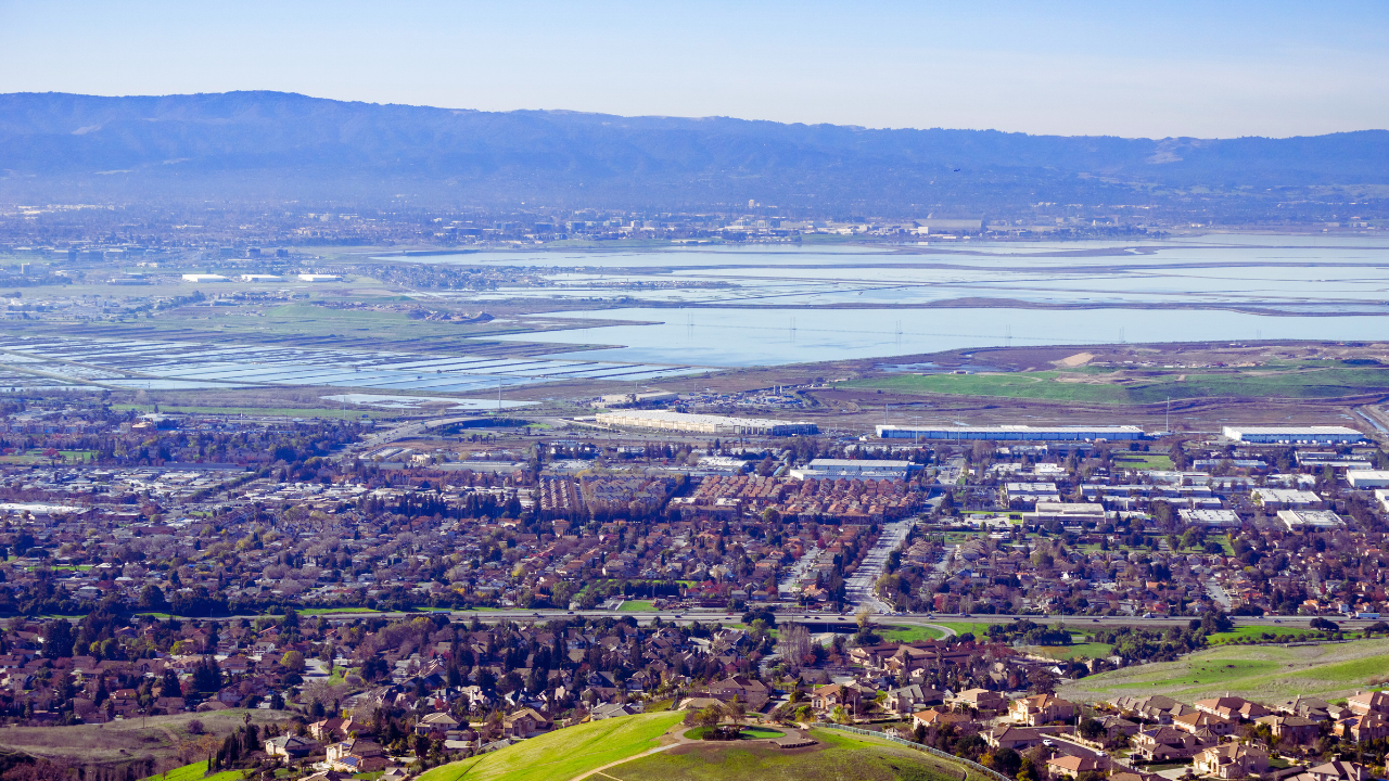 Wide city and valley view under clear blue sky, with a shimmering lake and mountains in the distance.
