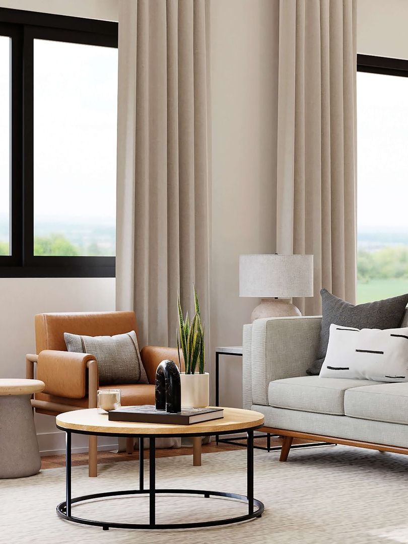 Modern living room with beige curtains, a gray sofa, tan chair, and round coffee table beside large windows