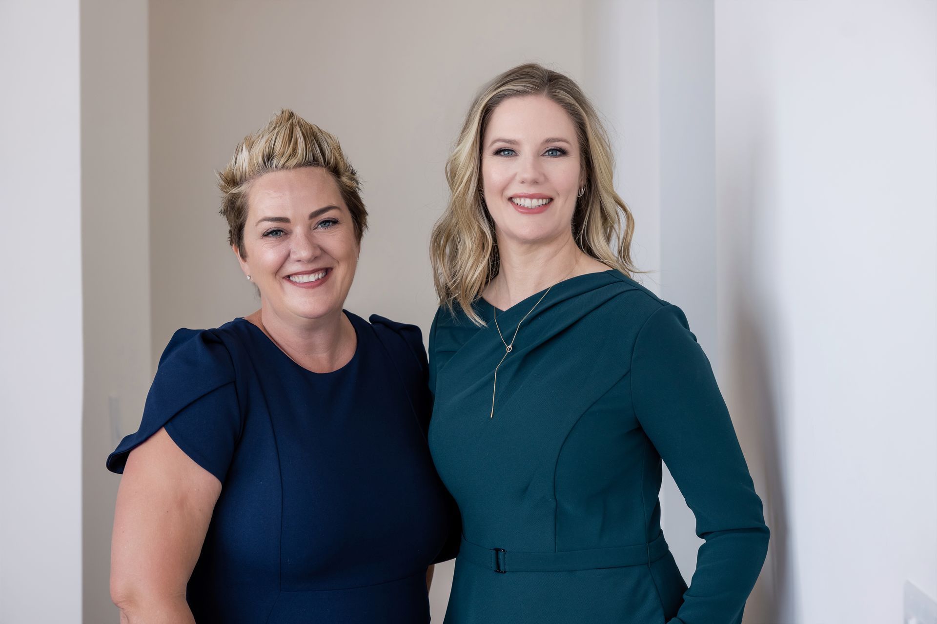 Two women standing indoors, smiling, in navy and teal dresses with a white wall background