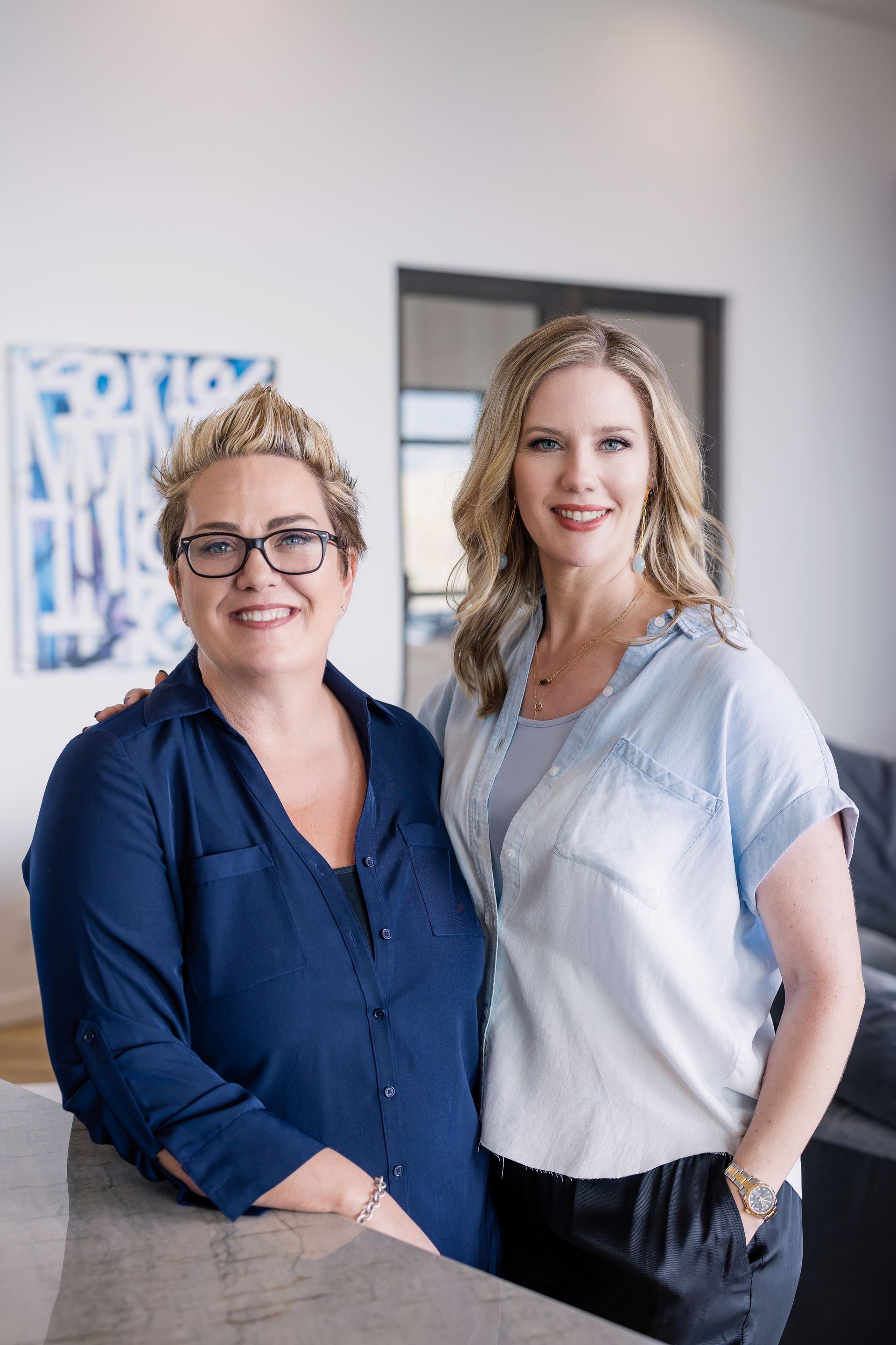 Two people standing indoors and smiling, wearing blue and white shirts in a bright office setting.