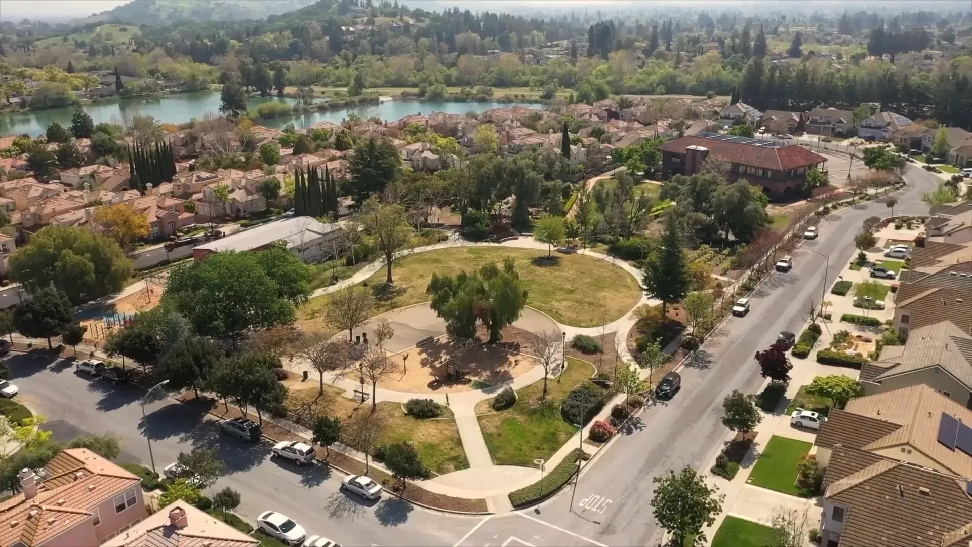 Aerial view of a suburban park with paths, trees, open lawns, and surrounding homes along a street.
