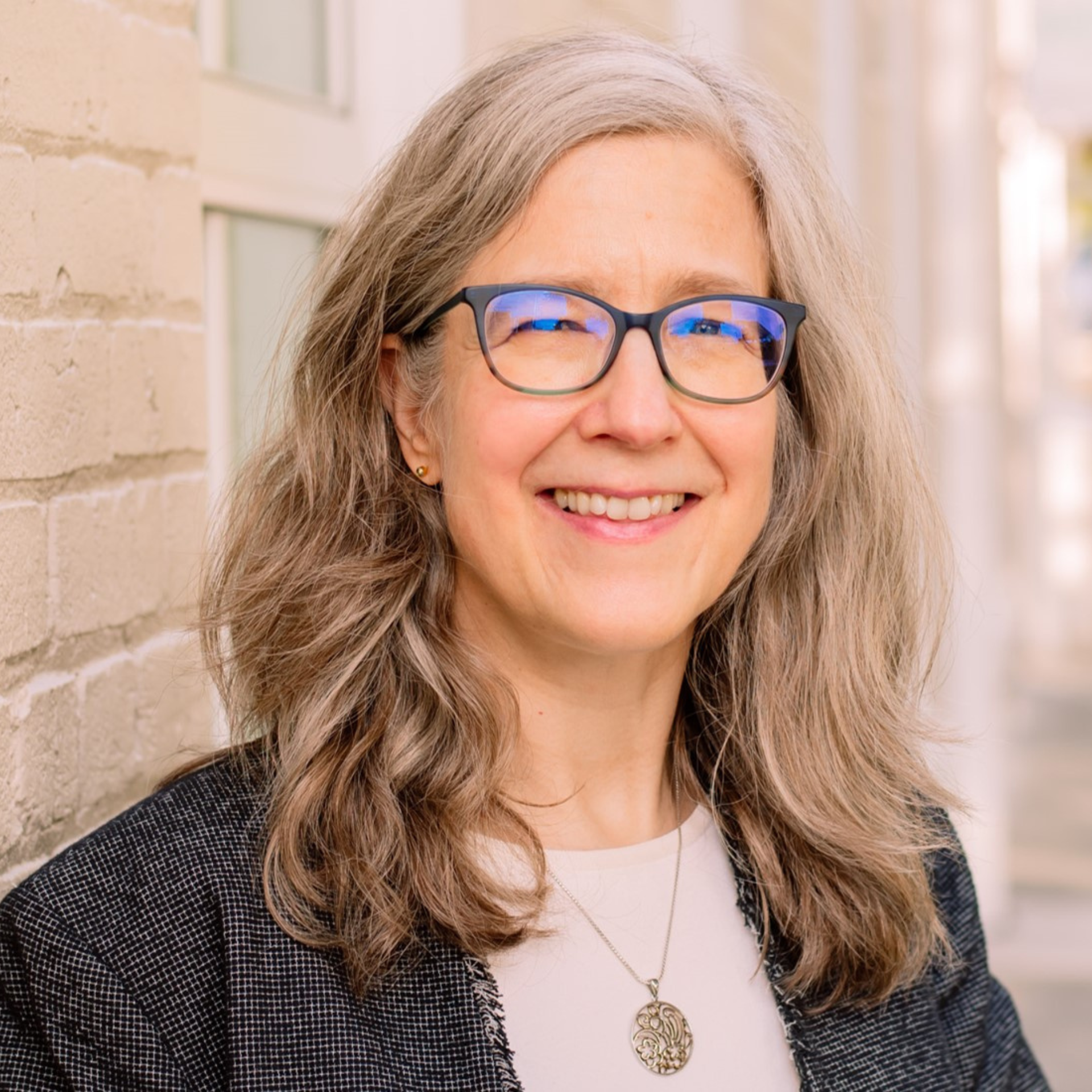 A woman wearing glasses and a necklace is smiling in front of a brick wall.