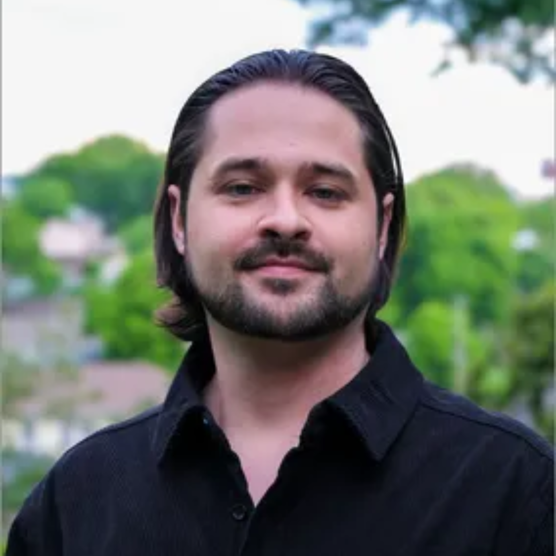 A man with long hair and a beard is smiling in the woods.