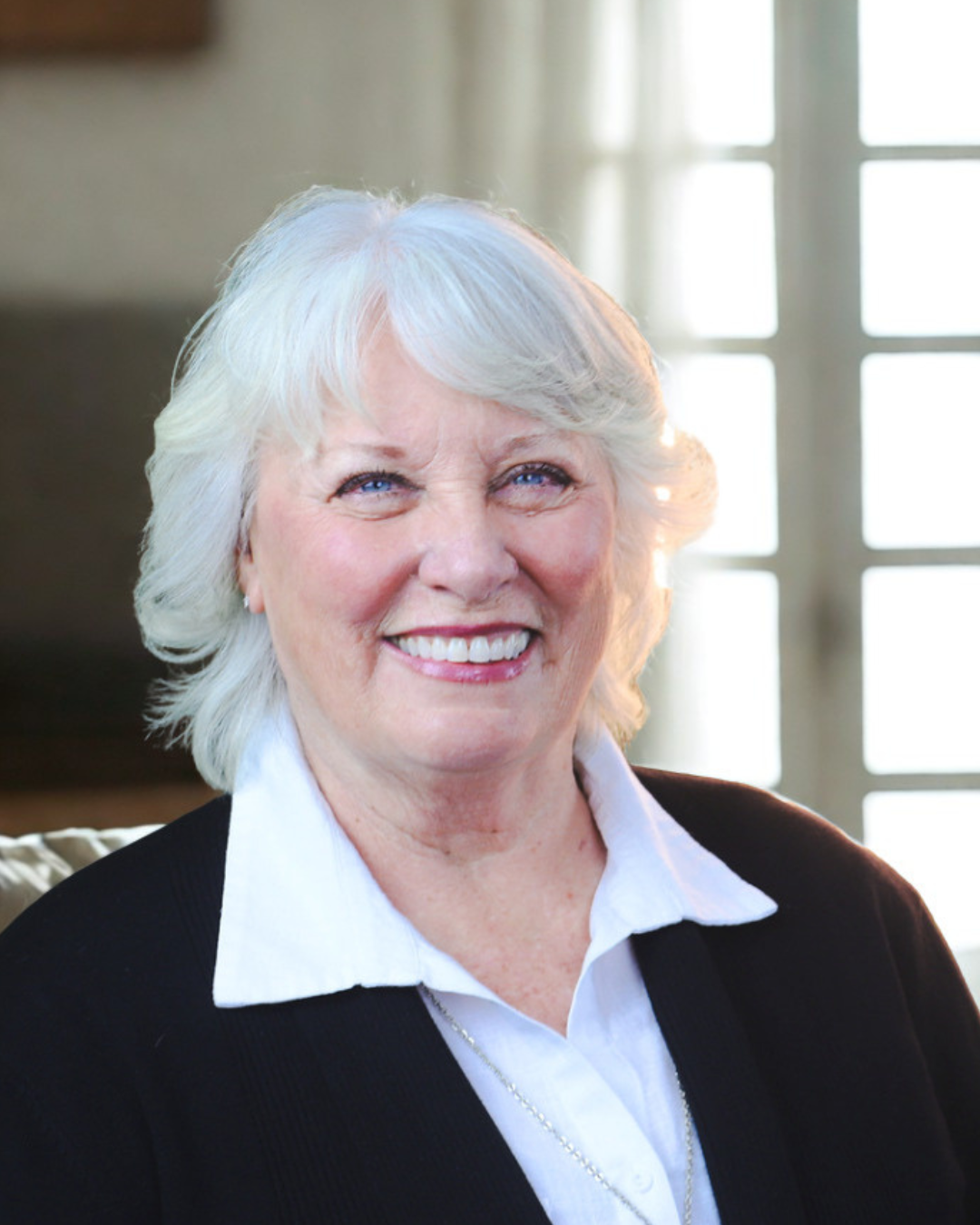 Smiling woman in a black blazer and white shirt, standing before bright windows.