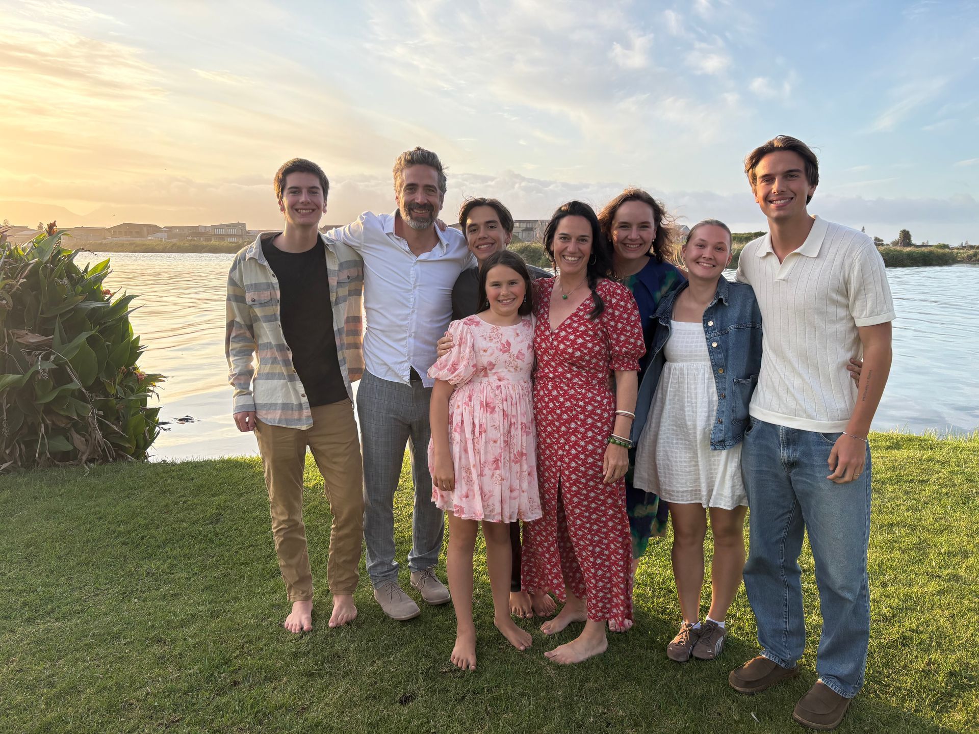 Six people smiling on a grassy lakeshore at sunset, with water and sky behind them.