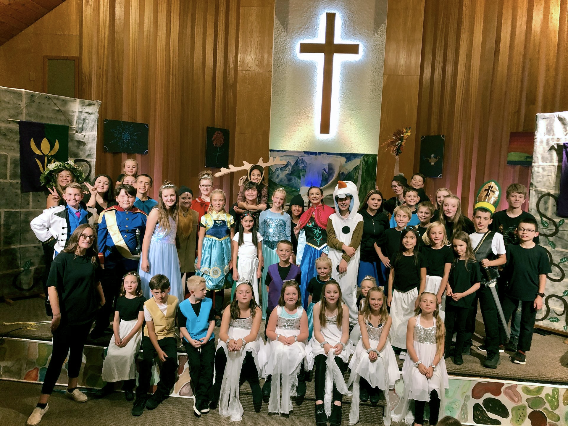 Large group posing on a church stage beneath a glowing cross, with wood-paneled walls and flags behind them