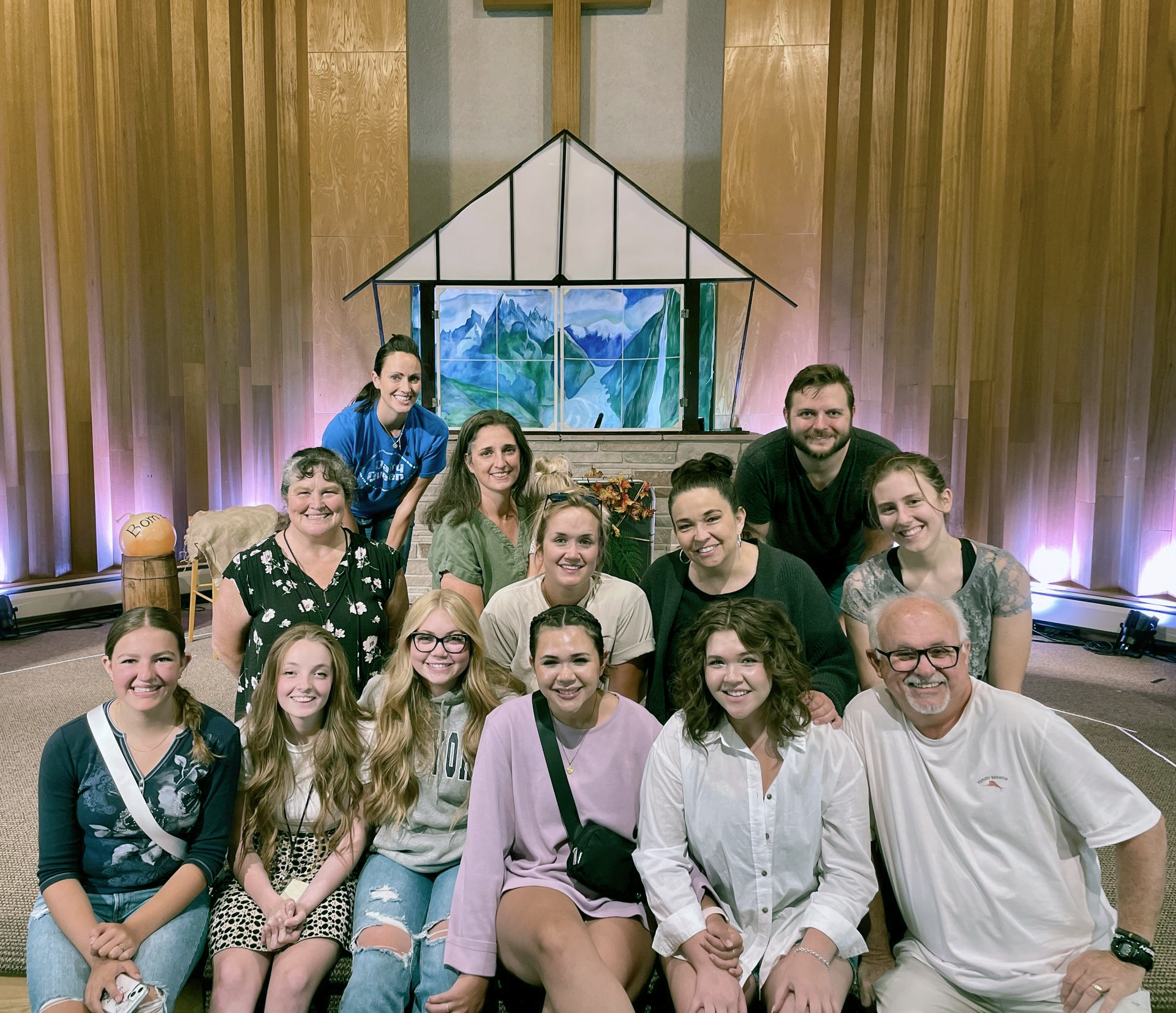 Group of people posing in a church sanctuary before a stained-glass display and wooden cross.