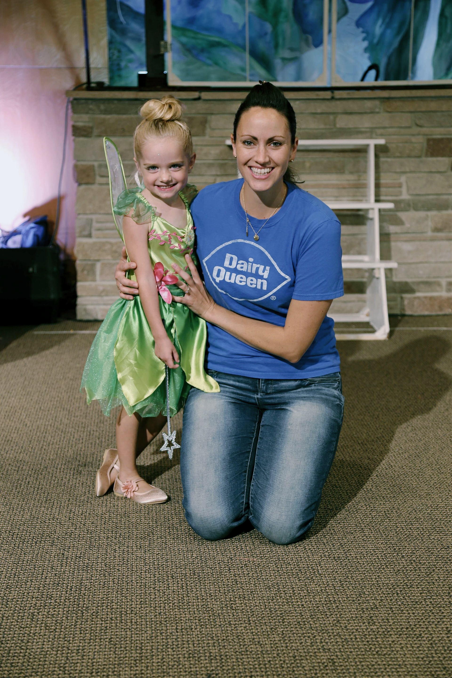 Woman kneeling beside a smiling girl in a green costume on a carpeted stage