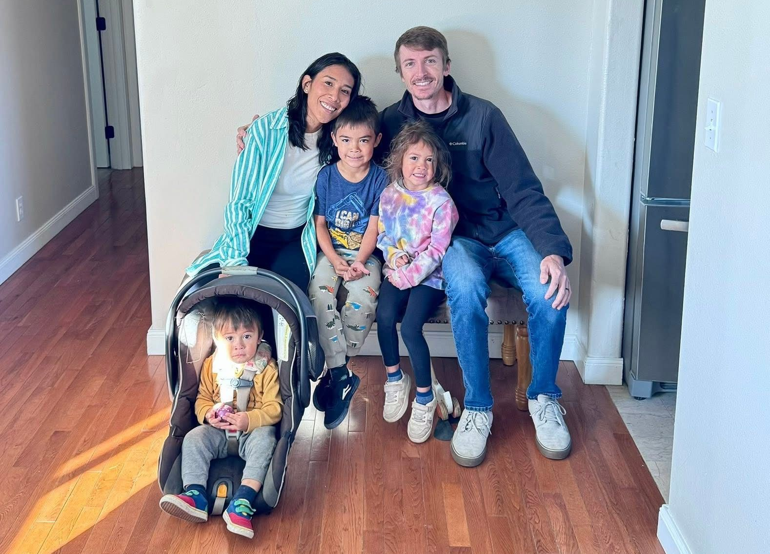 Family seated on a hallway floor, smiling beside a baby stroller with a child in it.