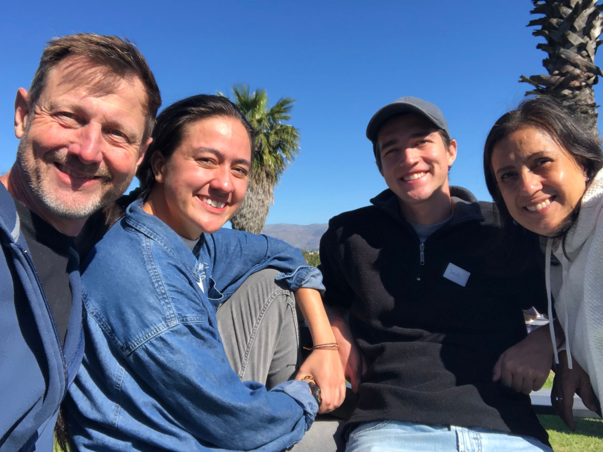 Four people smiling outdoors on a sunny day, seated together with blue sky and palm trees behind them