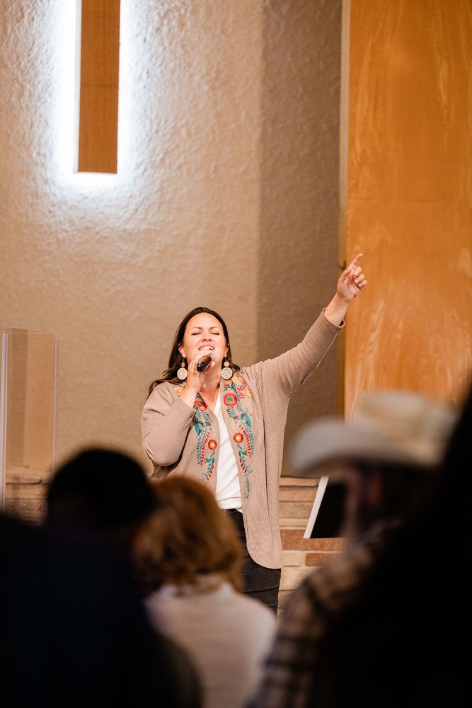 Speaker on stage gesturing upward, addressing an audience in a warmly lit room.
