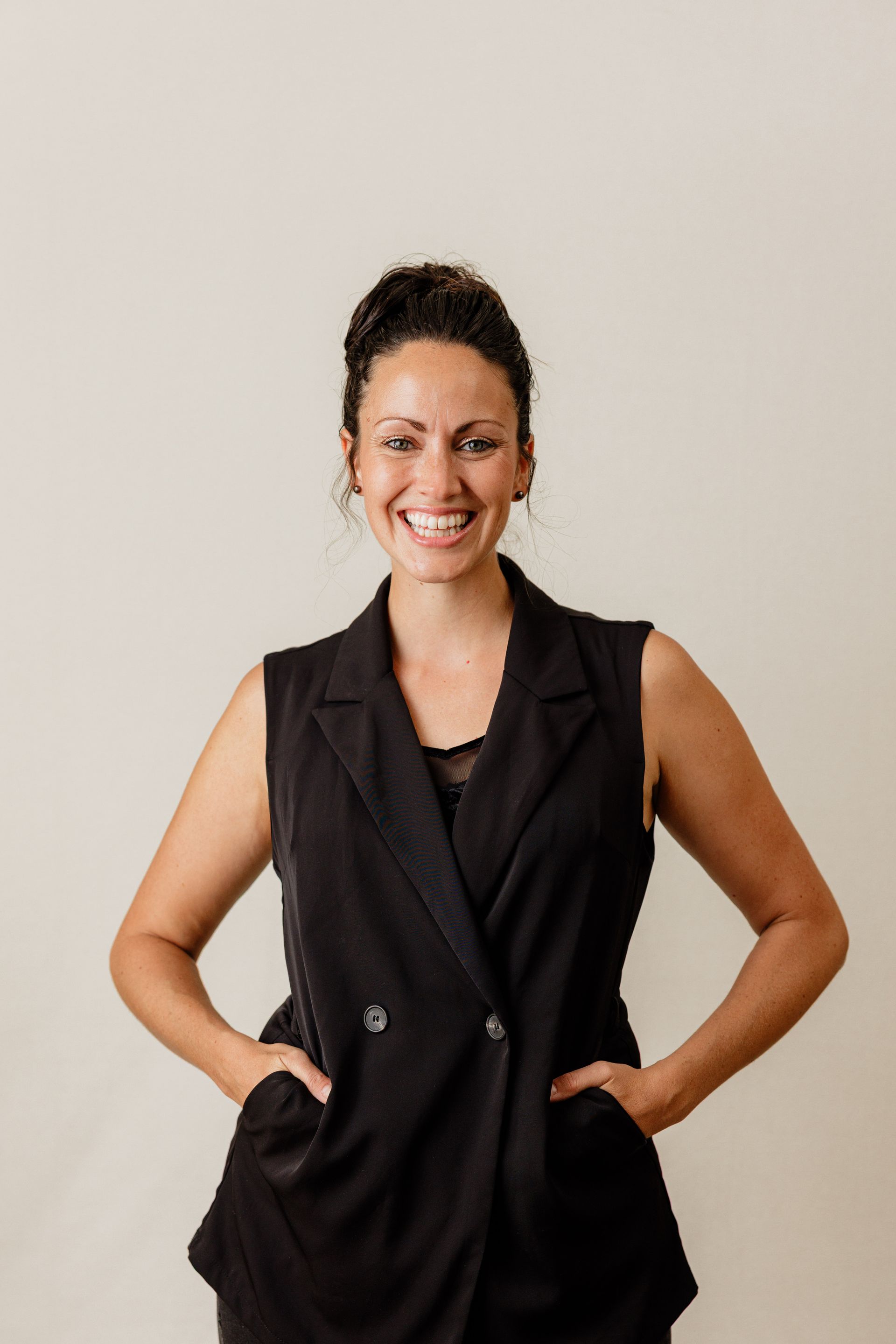 Smiling woman in black shirt and blue jeans, standing with hands in pockets against a plain background