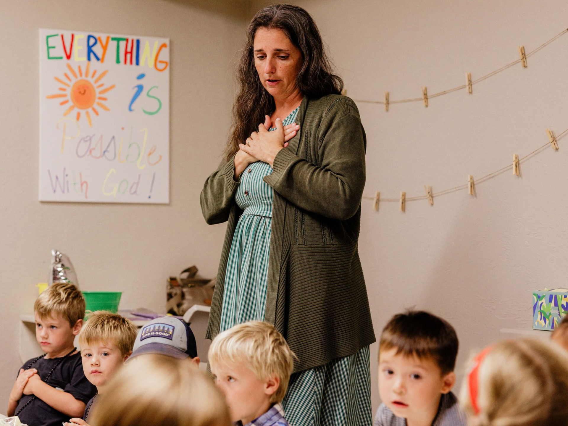 Teacher standing with hand on chest, speaking to young children in a classroom decorated with a poster and string lights.