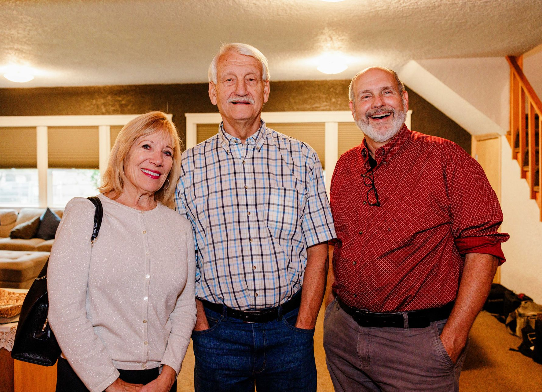 Three smiling people standing indoors in a bright living room, wearing casual shirts.