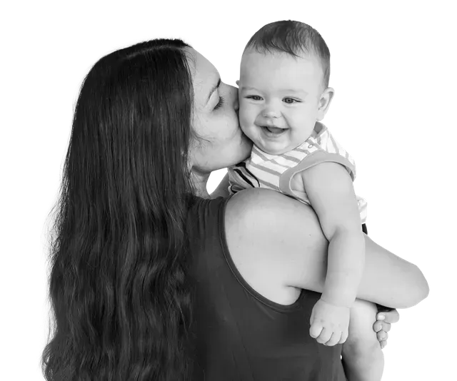 Woman kissing a smiling baby on the cheek; they are both in front of a white background.