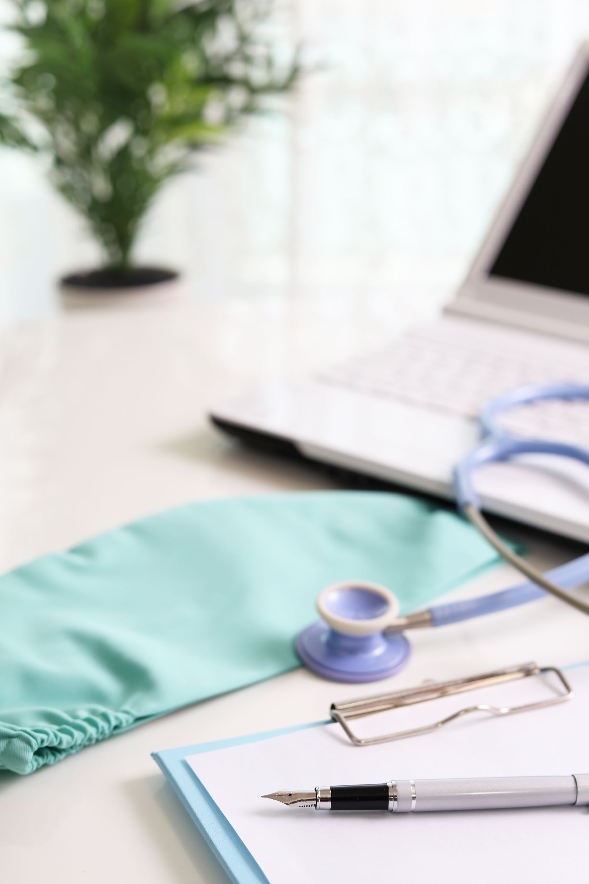 A doctor's desk: stethoscope, surgical cap, notepad with pen, laptop, and plant.