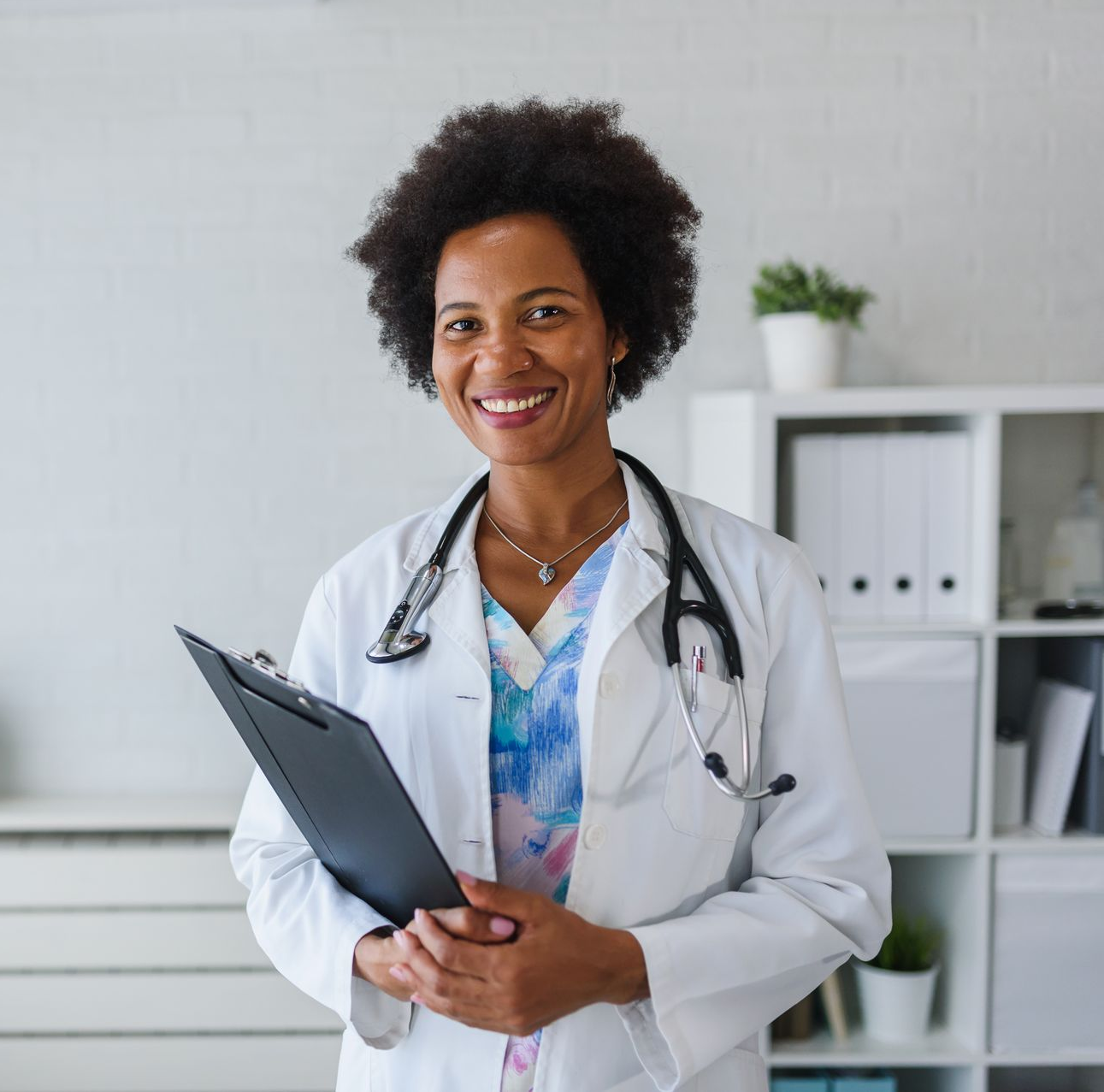 Doctor smiling, holding a clipboard, wearing a stethoscope, in a well-lit office.