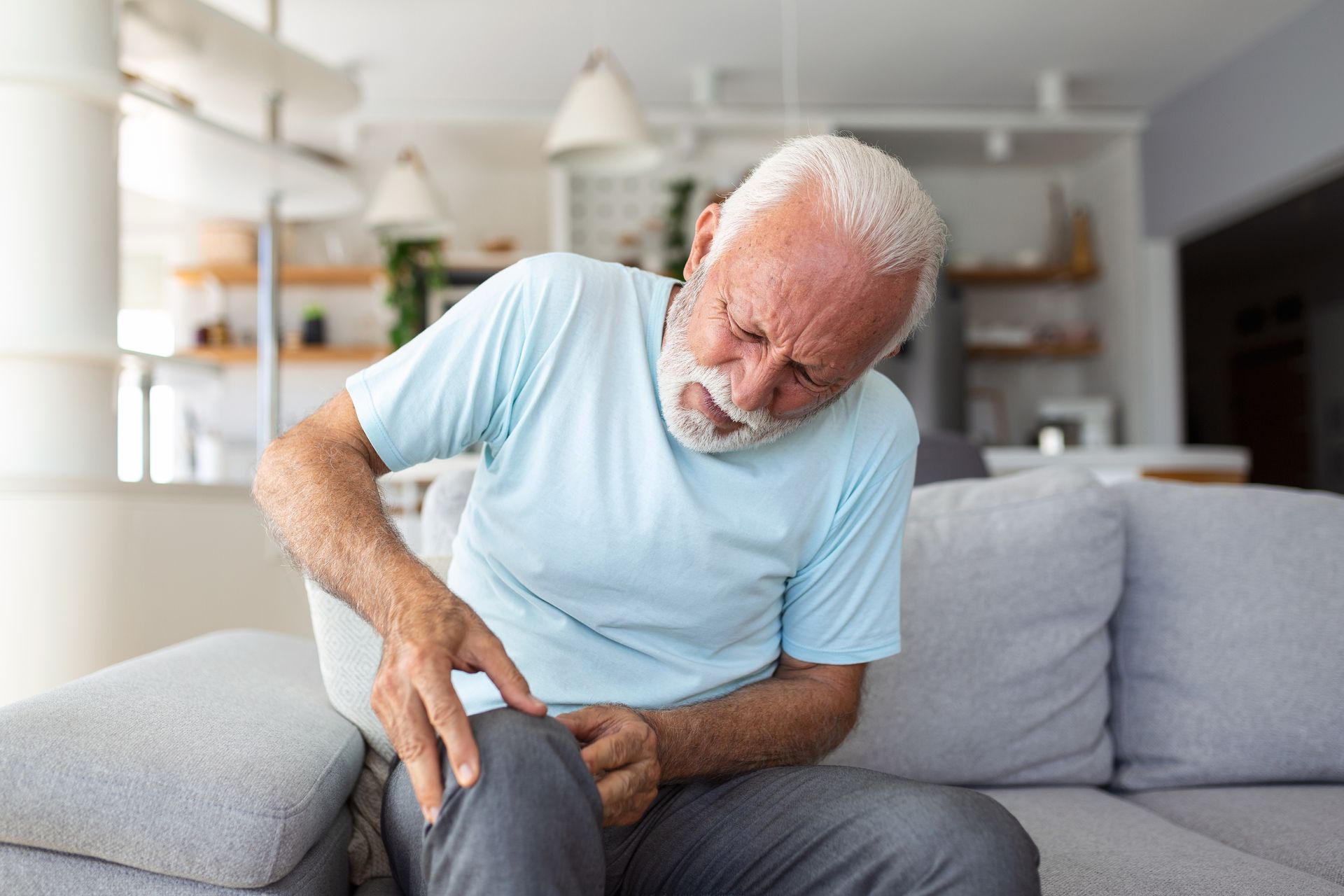 Older person on a sofa, grimacing and holding their knee in pain.