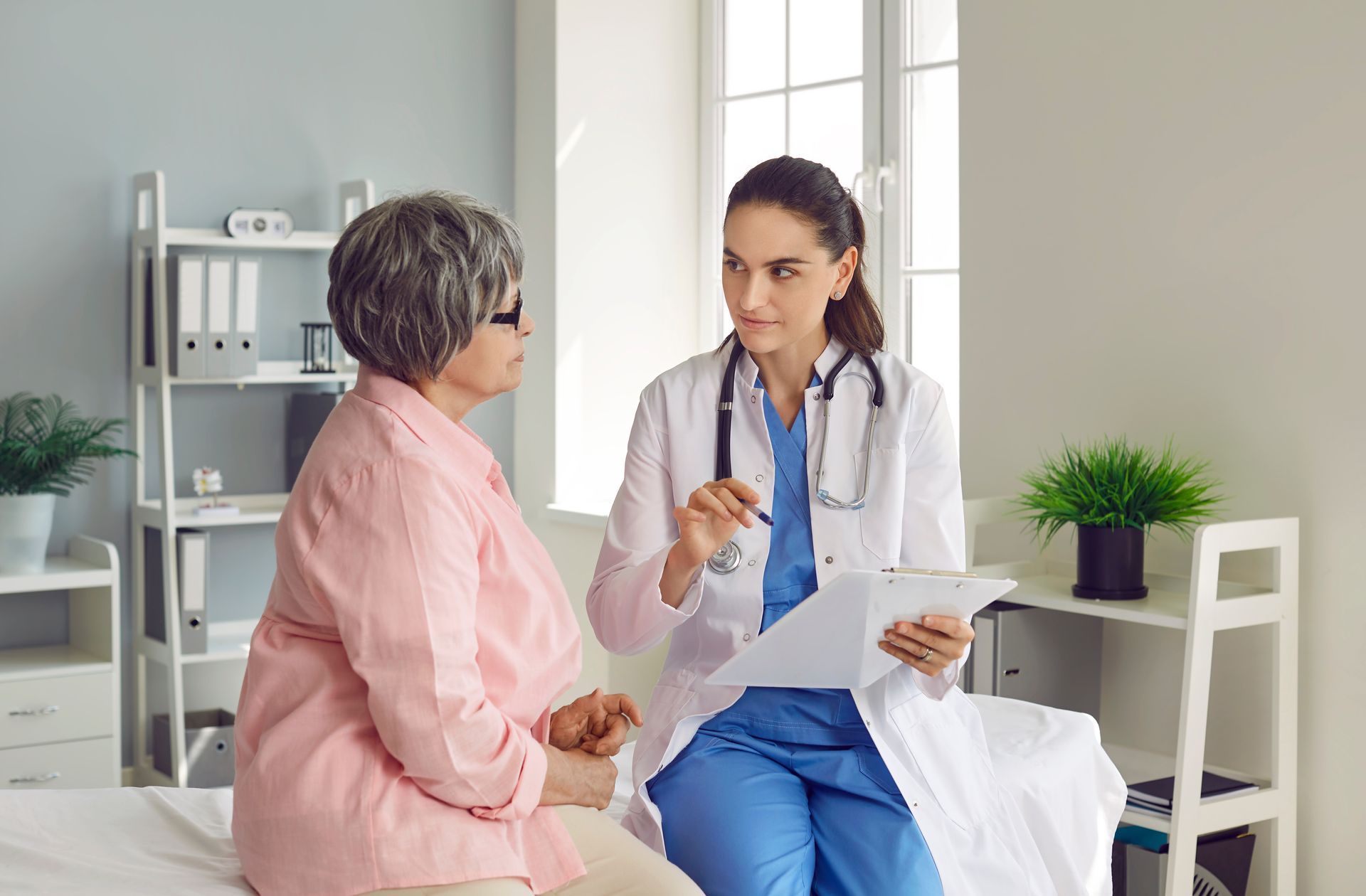 Doctor in white coat consults with a patient seated on a bed, holding a clipboard; indoor setting.