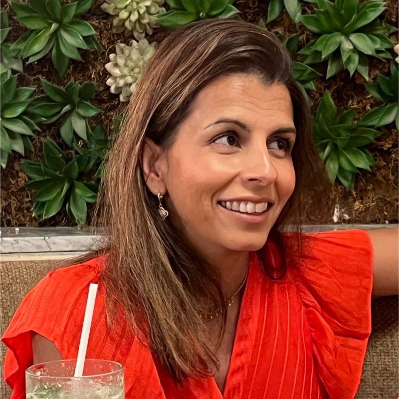 A woman wearing glasses and an orange shirt is standing with her arms crossed and smiling.