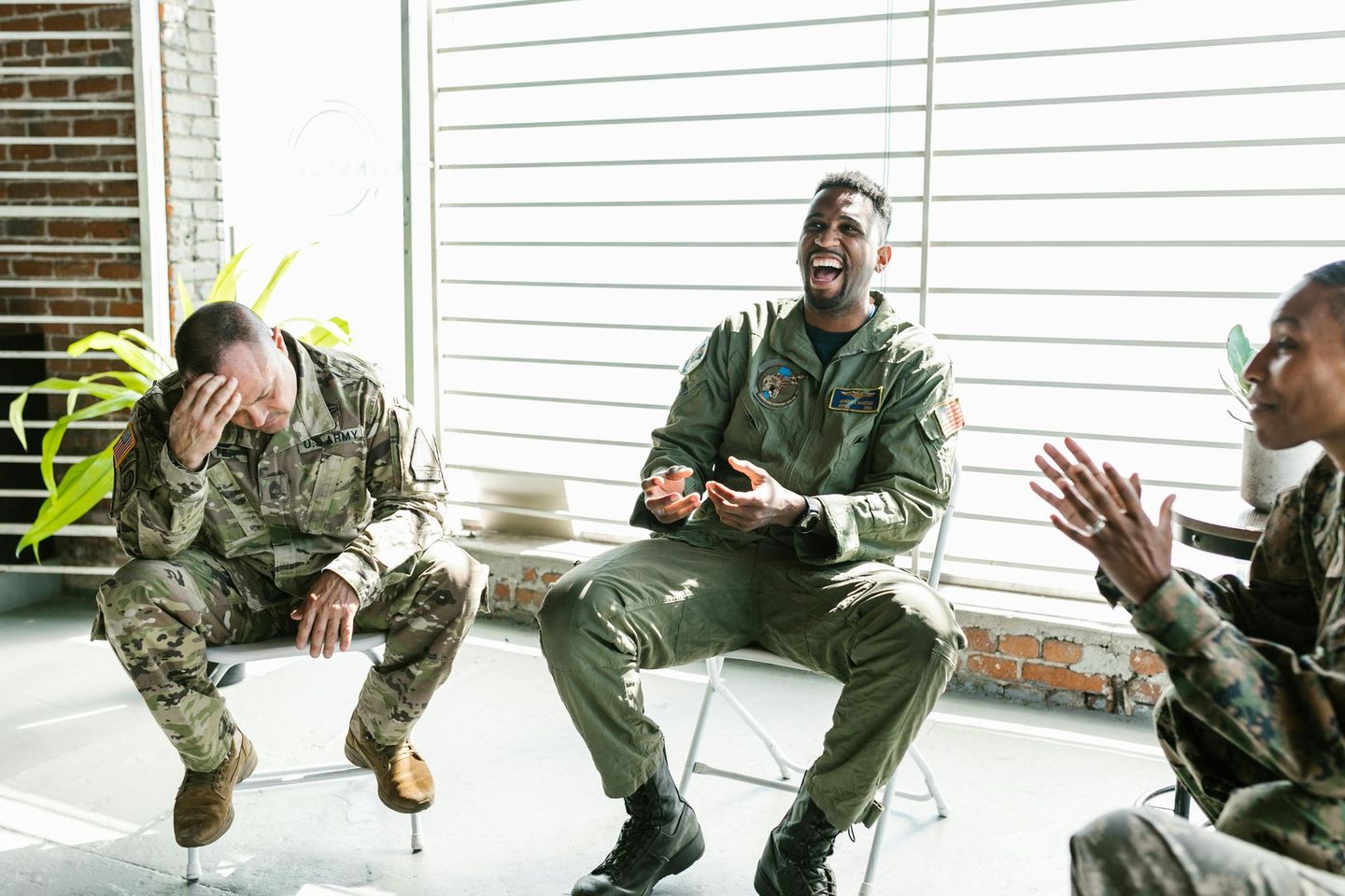 Three soldiers in camouflage uniforms sit and laugh in a bright room with white blinds.