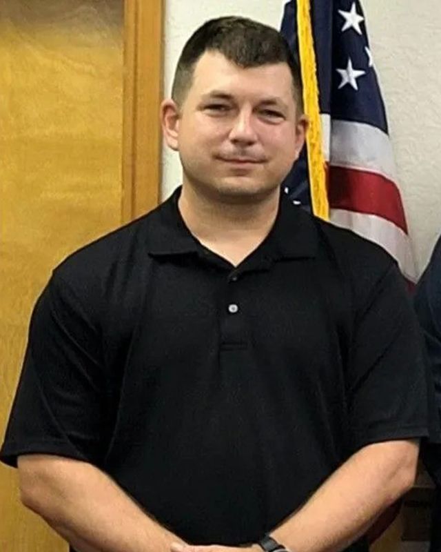A man in a black shirt is standing in front of an American flag.