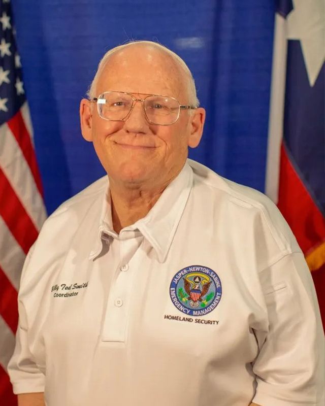 A man wearing glasses and a white shirt stands in front of American flags