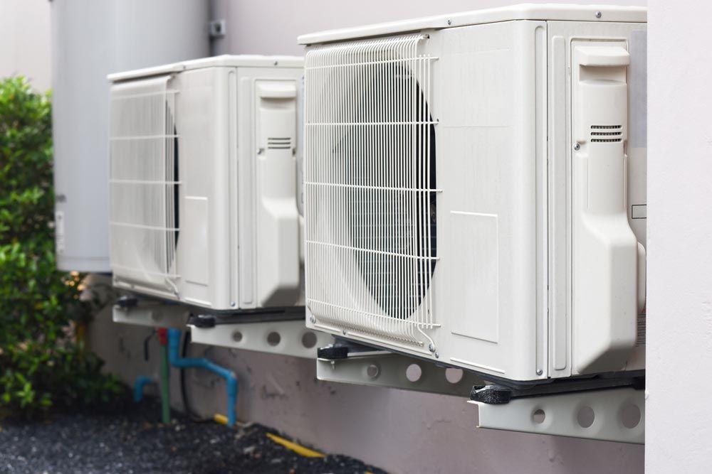 Air conditioning unit mounted on a brick wall, near a lawn and gutter, with a white pipe leading upward.