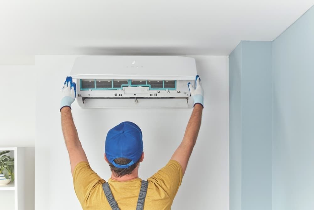 A person in a blue cap installing a white AC unit on a wall, indoors.