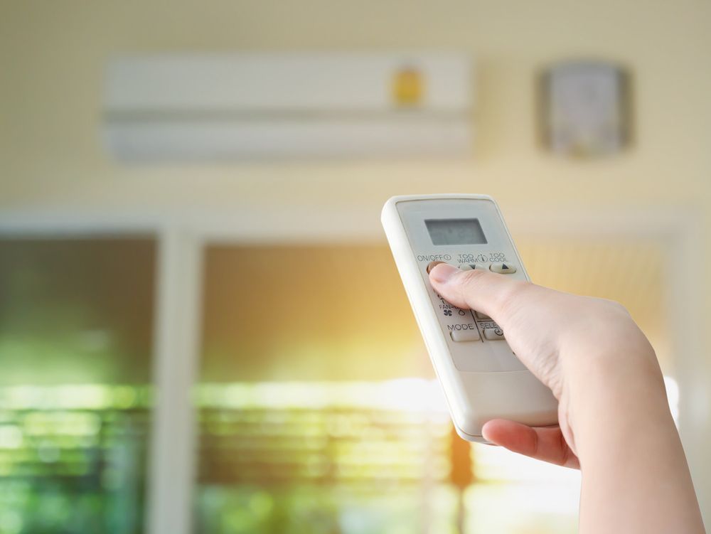 Hand holding an AC remote, pointing at the wall-mounted unit, with a window in the background.