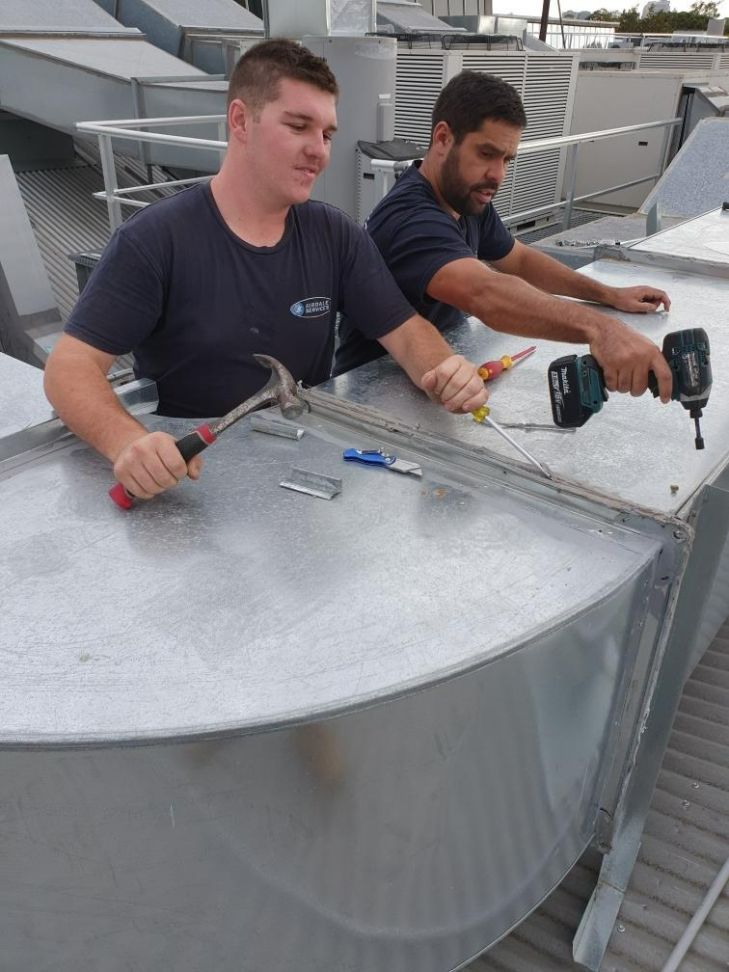 Two men working on a metal structure on a rooftop. One uses a hammer, the other a drill.