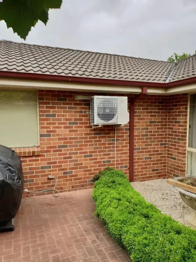 Air conditioning unit mounted on a red brick exterior wall under a tiled roof. A green hedge is below.