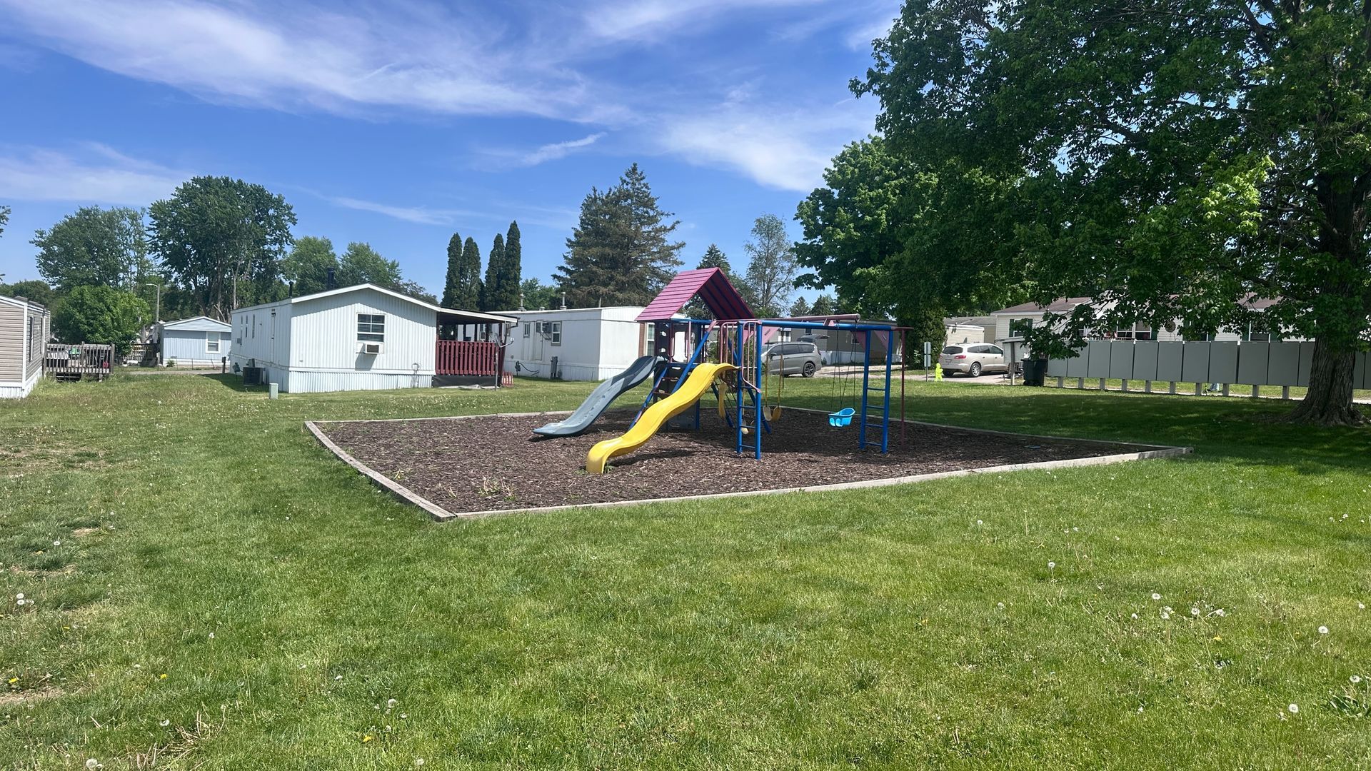 Playground with a yellow slide and blue climbing structure on mulch, surrounded by grassy lawn and trees