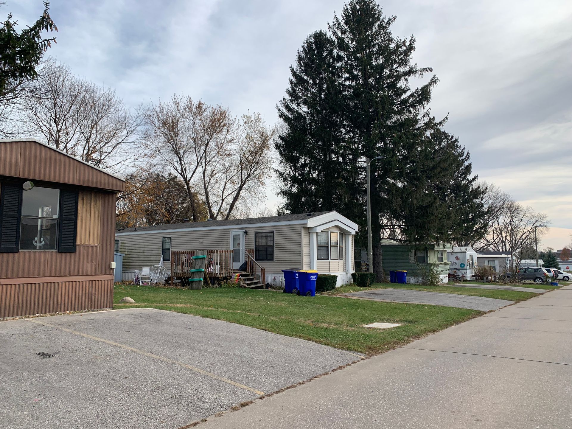Mobile home park with a gray trailer, blue bins, and tall trees beside a paved road