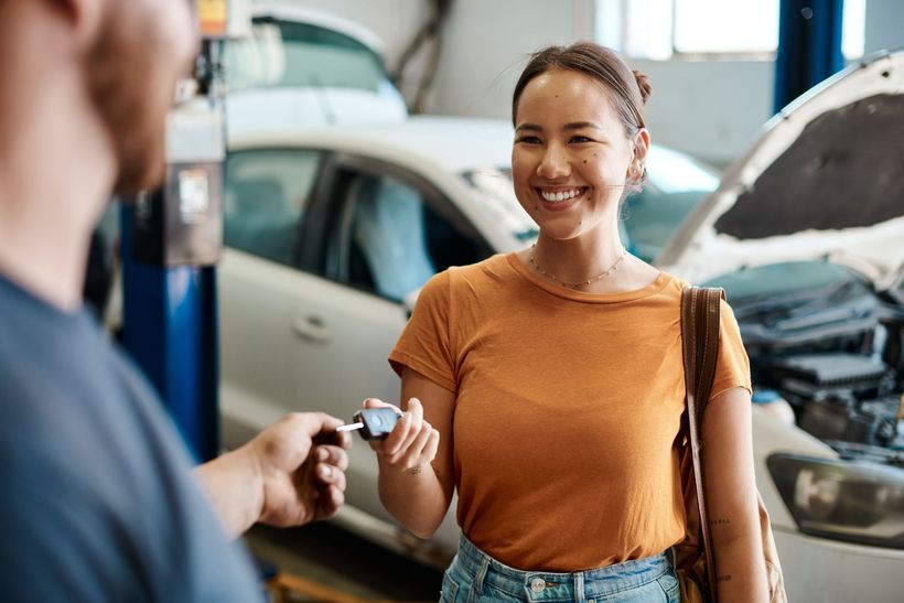 Woman smiling at a car repair shop, holding keys near a car with its hood open.