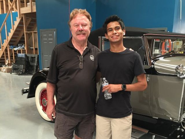 Two men stand smiling beside a vintage car in a garage with blue walls.