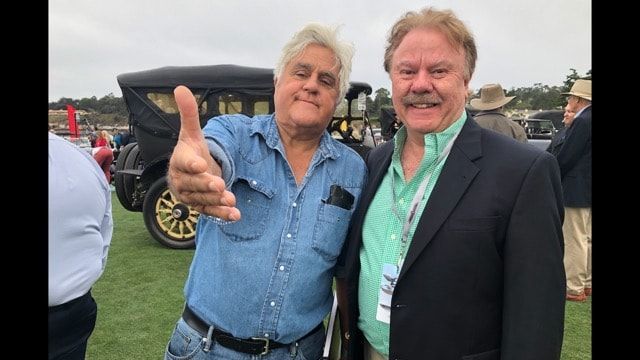 Two men smiling outdoors, one giving a thumbs-up in front of a vintage car display.