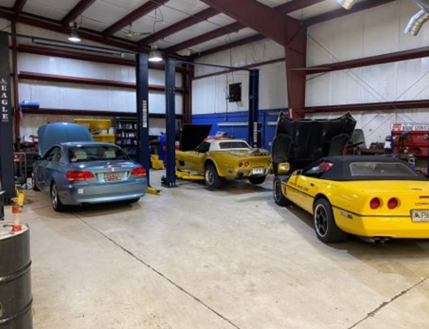 Three classic cars in a warehouse garage, including a blue sedan and two yellow sports cars.