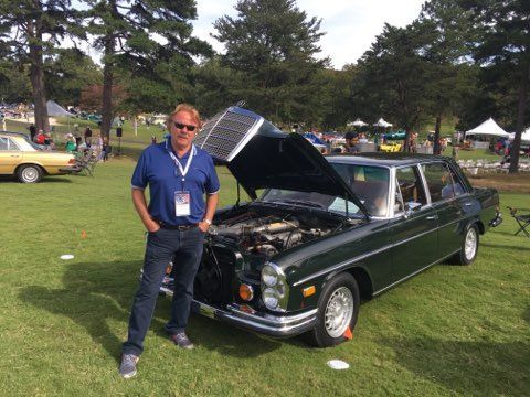 Man standing beside a dark green classic car with its hood open at an outdoor car show